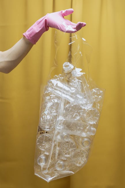 A close-up view of a large black plastic rubbish bag, tightly tied at the top, resting against a plain, light-colored wall. The bag appears full, with noticeable creases and glossy surface texture ref